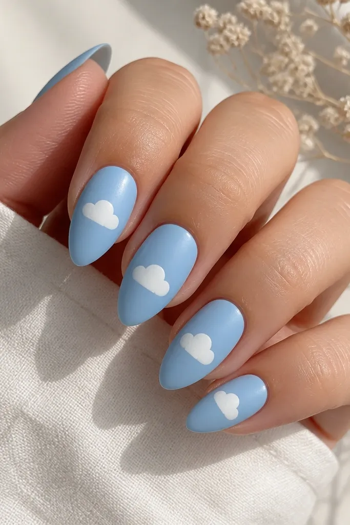 Short stiletto nails painted sky blue in a satin finish. The tips have small white cloud shapes, like tiny puffs, centered on each nail. The overall look is matte-soft with crisp white clouds.