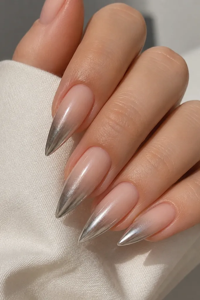 Short stiletto nails with a nude base. From mid-nail to the tip, there's a steel silver chrome fade that gets stronger toward the point. The rest stays soft nude and glossy.