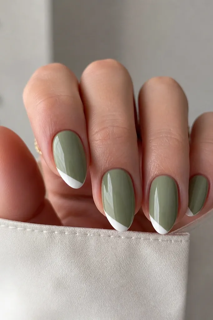 Short stiletto nails painted muted sage green glossy. The tips have a white French border but it's slightly angled, like a subtle diagonal French. The point stays short and clean.