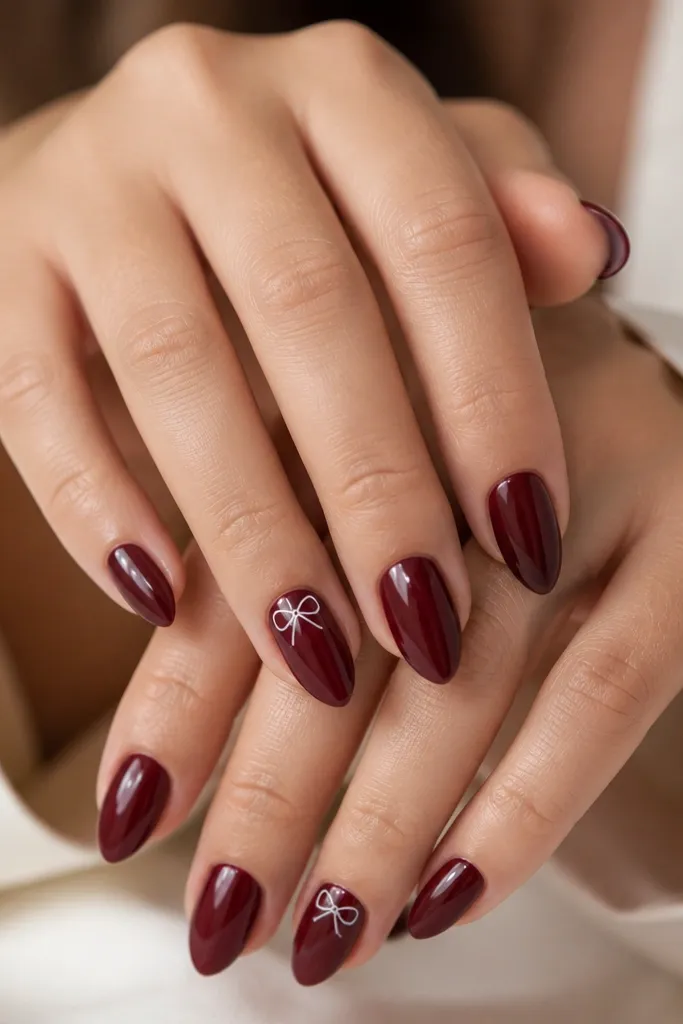 Short stiletto nails painted glossy burgundy wine. On the ring finger nails, there's a tiny white bow with two small loops and a center knot placed near the upper third. Other nails are plain burgundy.