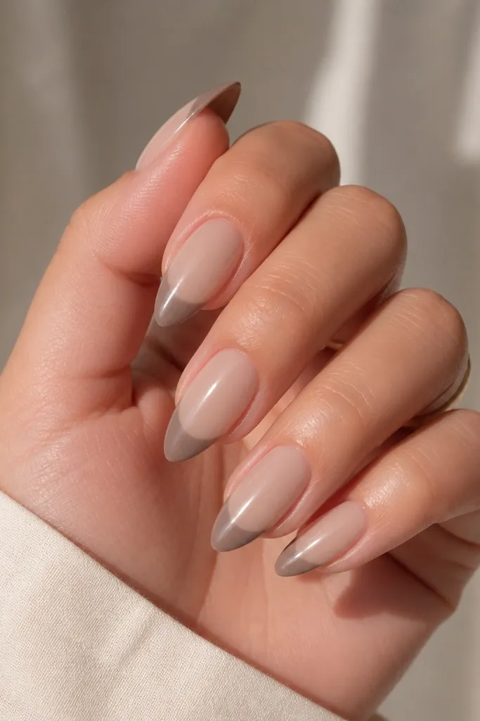 Short stiletto nails in sand beige nude glossy. Each nail has a taupe glossy tip that varies slightly in thickness, like a skittle effect. The taupe tips blend softly into the nude base.
