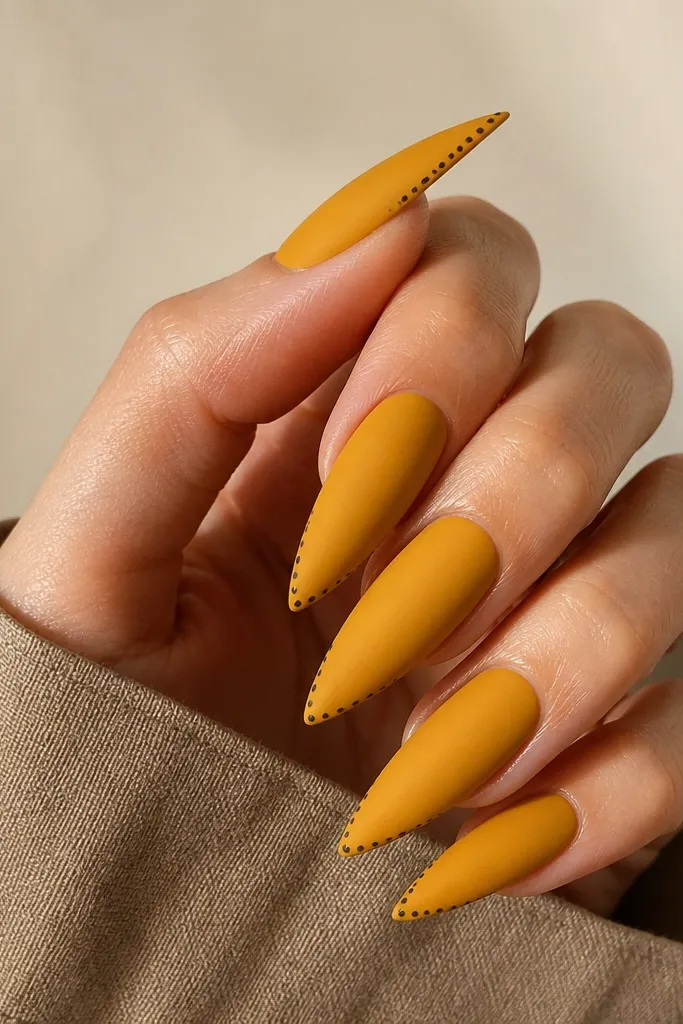 Stiletto nails in mustard yellow with a matte finish. A micro-French line runs across the tip in chocolate-brown dots, with dots only on the outer edge of the nail. The rest of the nail stays solid mustard.