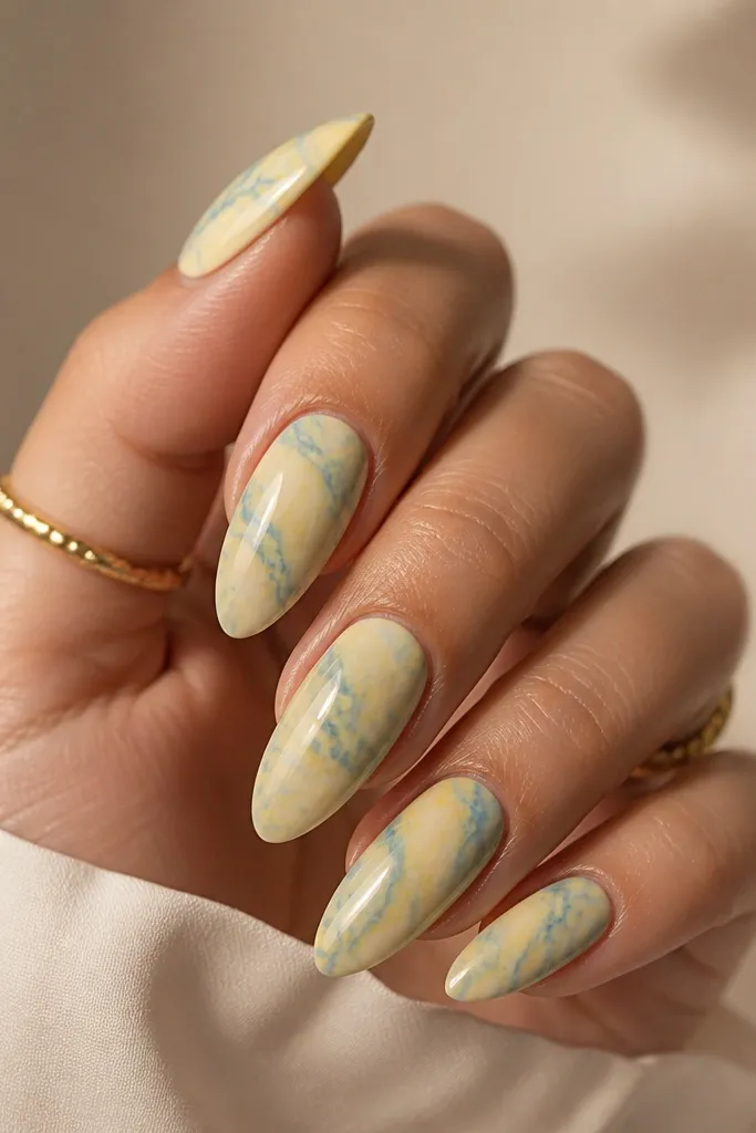 Marble effect nails with butter-yellow base and soft white-and-light-blue swirls like stone. Glossy top coat with smooth finish.